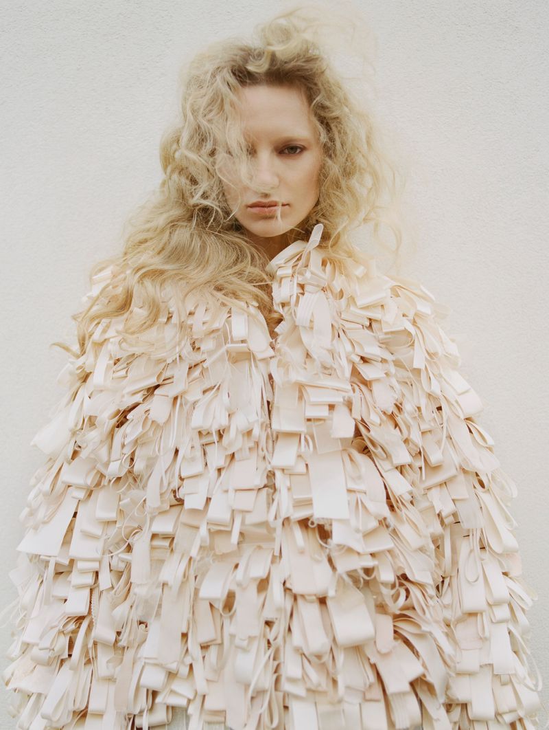 Blonde woman poses in a ribbon-covered jacket against a white background in a fashion editorial shot with soft lighting.