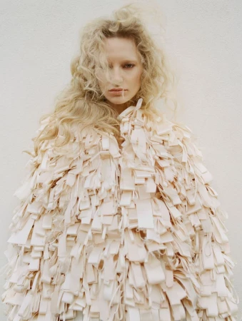 Blonde woman poses in a ribbon-covered jacket against a white background in a fashion editorial shot with soft lighting.