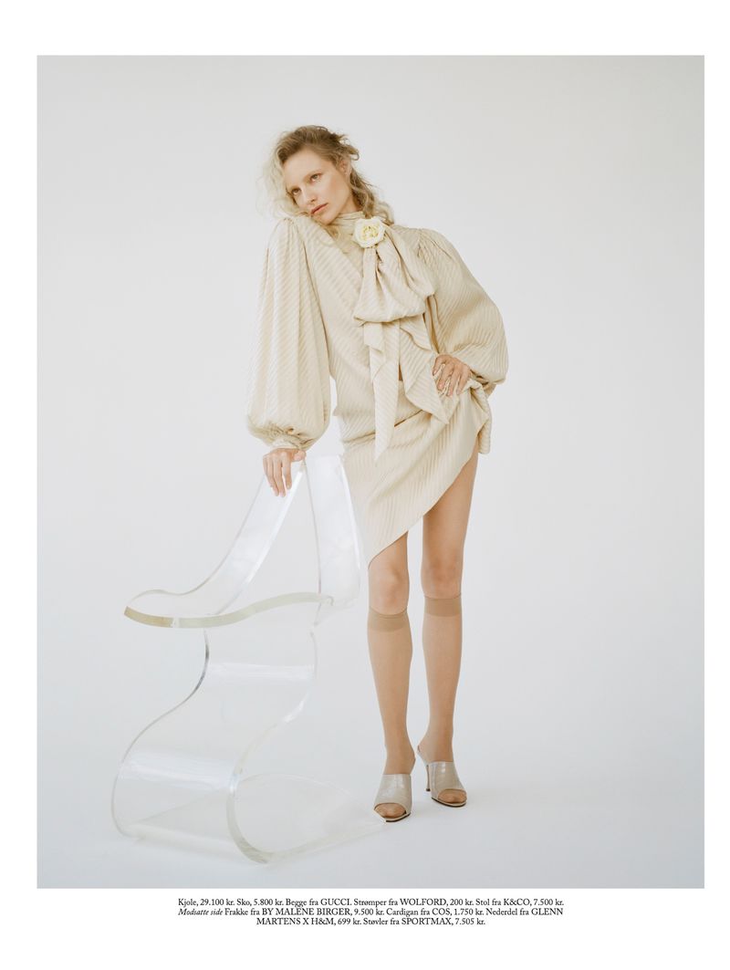 Fashion shot of woman in beige dress with oversized bow and blonde hair posing next to clear acrylic chair against white background.