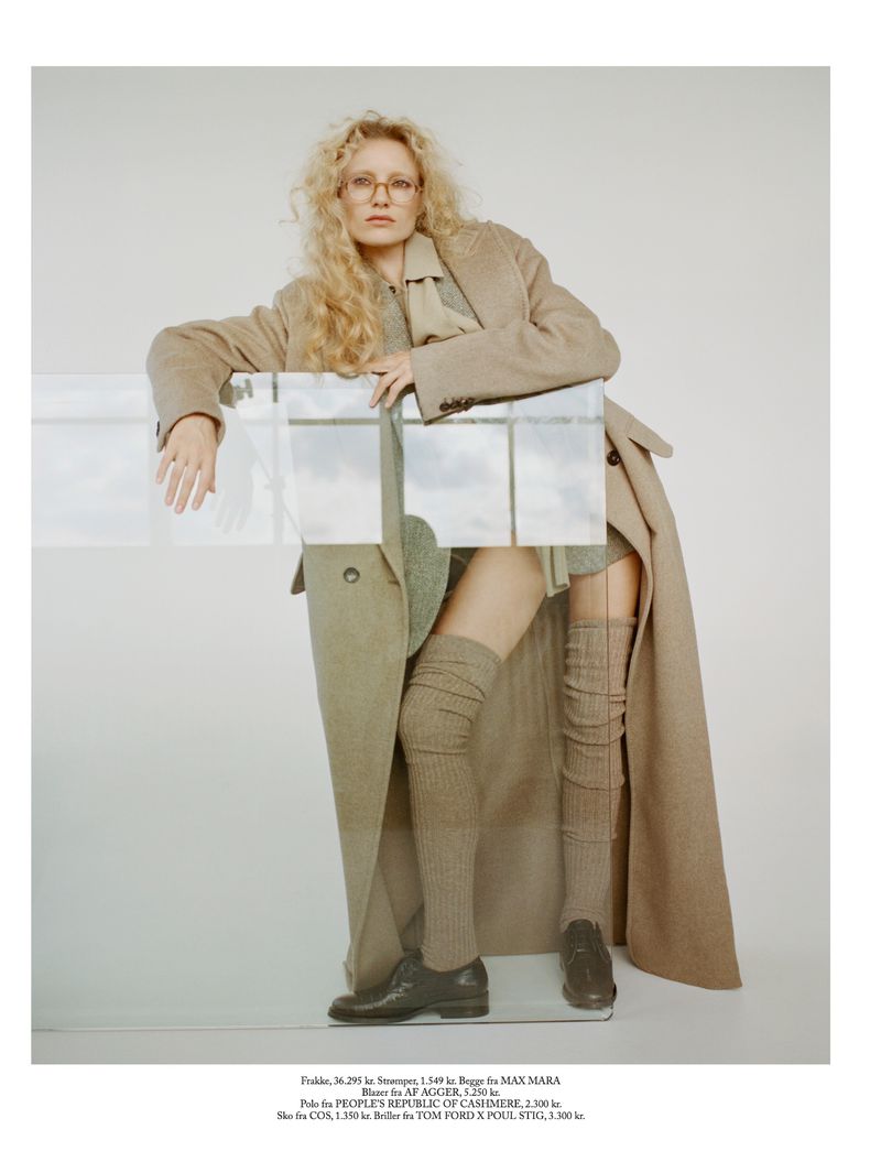 A woman with blonde curly hair poses in a tan trench coat, dress, and hosiery in a studio with a glass block prop.