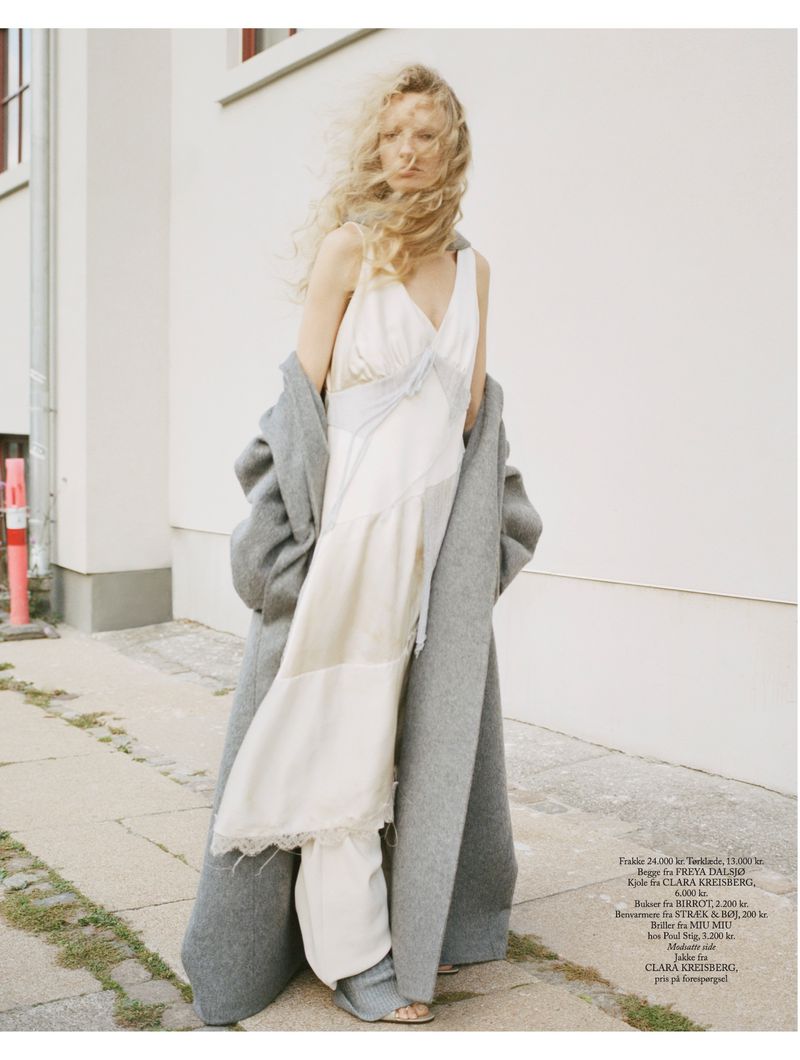 Woman wearing a white dress and a gray coat standing on a city street, her long blonde hair blowing in the wind.