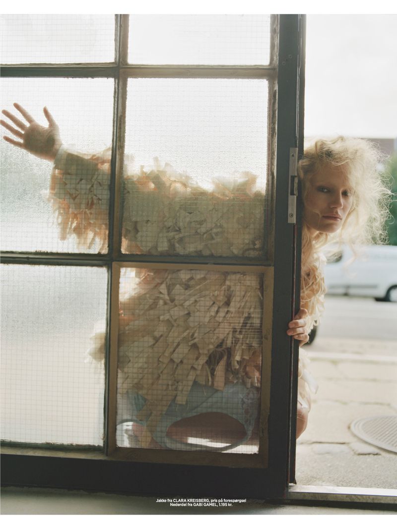 Woman with blonde curly hair poses behind a gridded window with a vintage van in the background on a city street.
