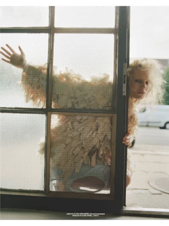 Woman with blonde curly hair poses behind a gridded window with a vintage van in the background on a city street.