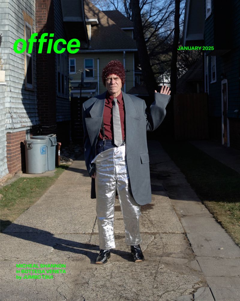 Adult male in silver pants and oversized gray suit jacket stands on a walkway, waving his left hand outside a house with a green lawn.
