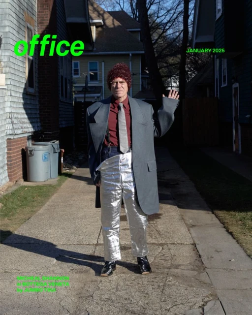 Adult male in silver pants and oversized gray suit jacket stands on a walkway, waving his left hand outside a house with a green lawn.