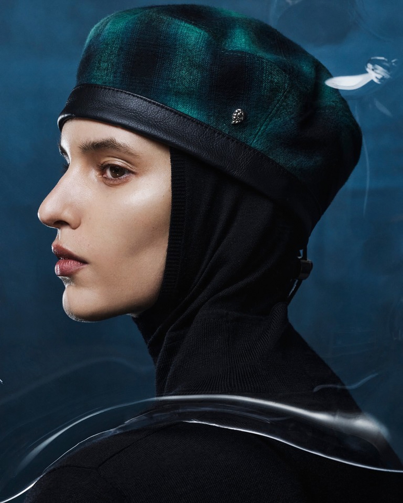 Profile view of a woman wearing a green plaid beret and black hooded top against a blue background with abstract light trails.