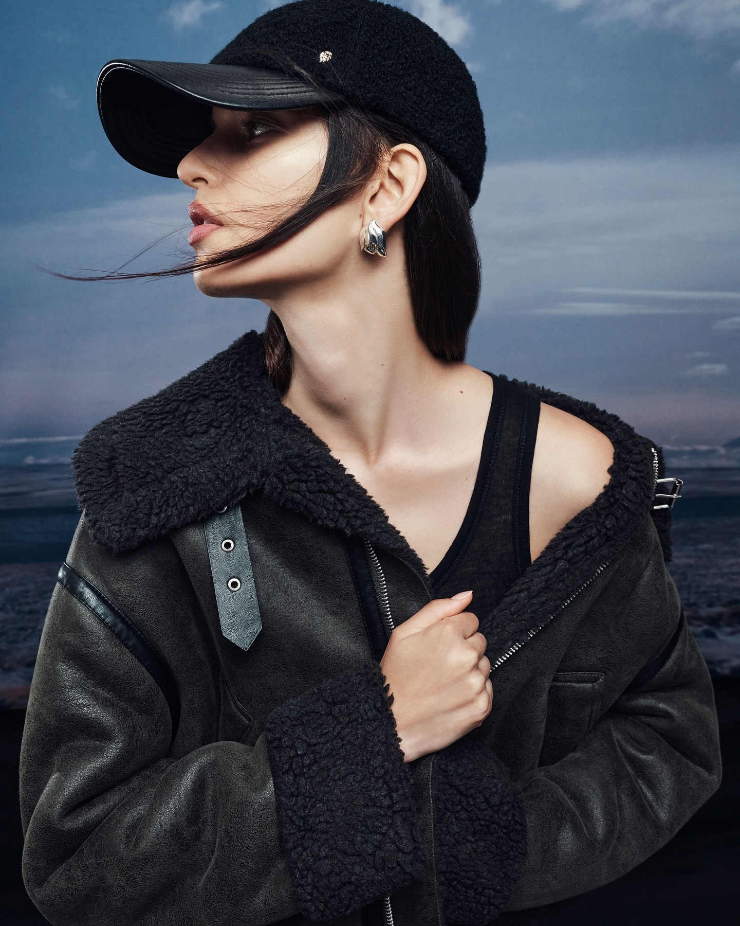 A woman in a black shearling jacket and cap looks off to the side against a blue and grey landscape background in a studio shot.