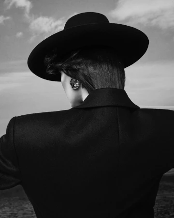 Monochrome fashion portrait of a model wearing a wide-brimmed black hat and tailored jacket against an open sky