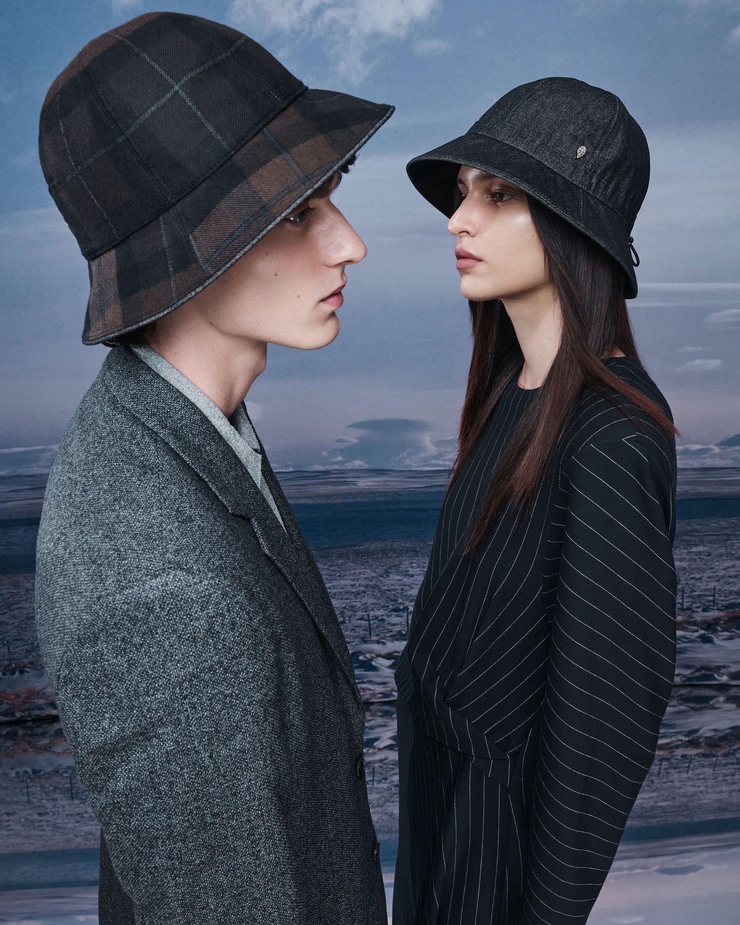 Two people wearing hats and coats stand in front of a blurred landscape with mountains and water in a fashion-style outdoor shot.
