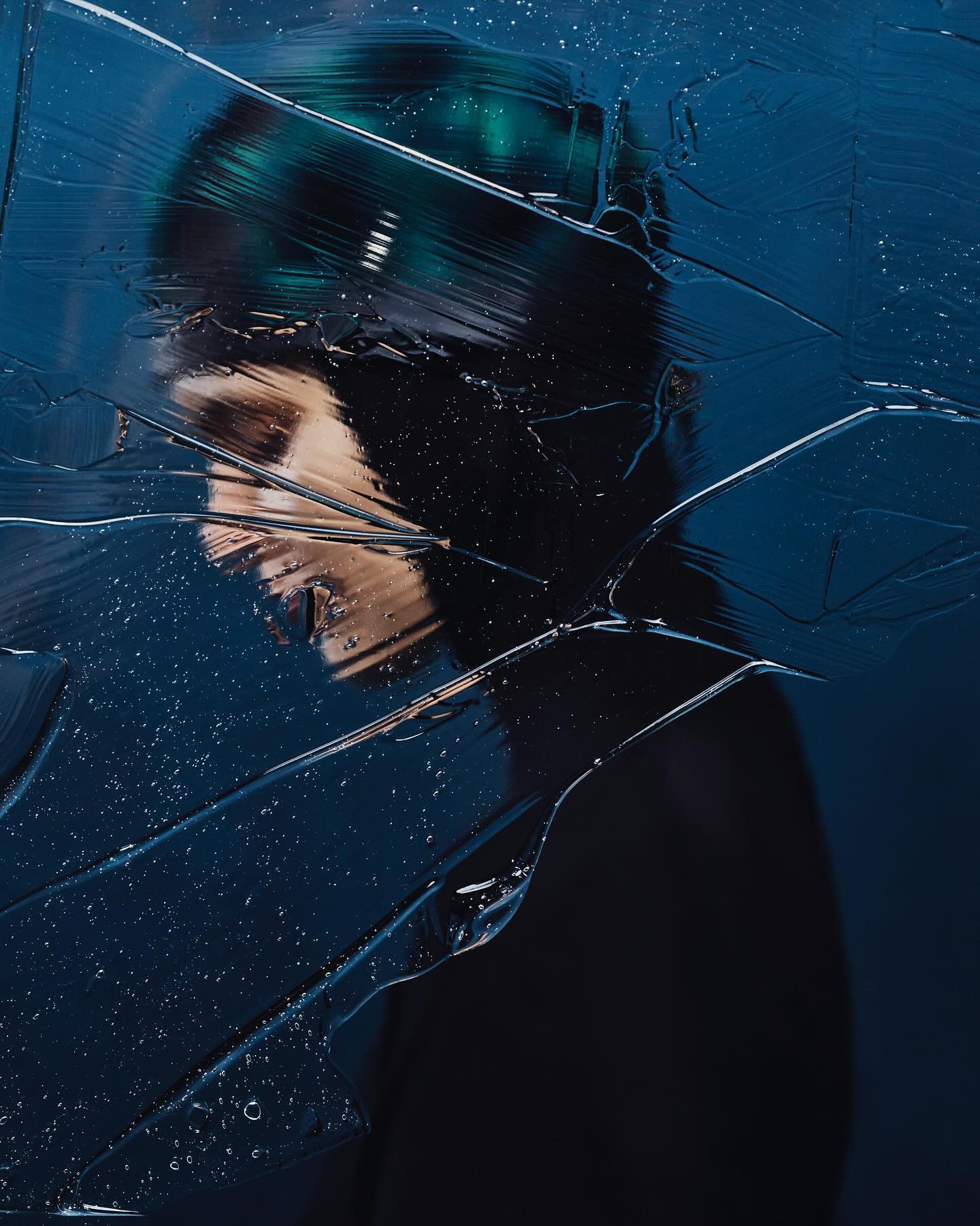 Surreal portrait of a person obscured by textured glass with water droplets in dark blue tones. Abstract and enigmatic photography.