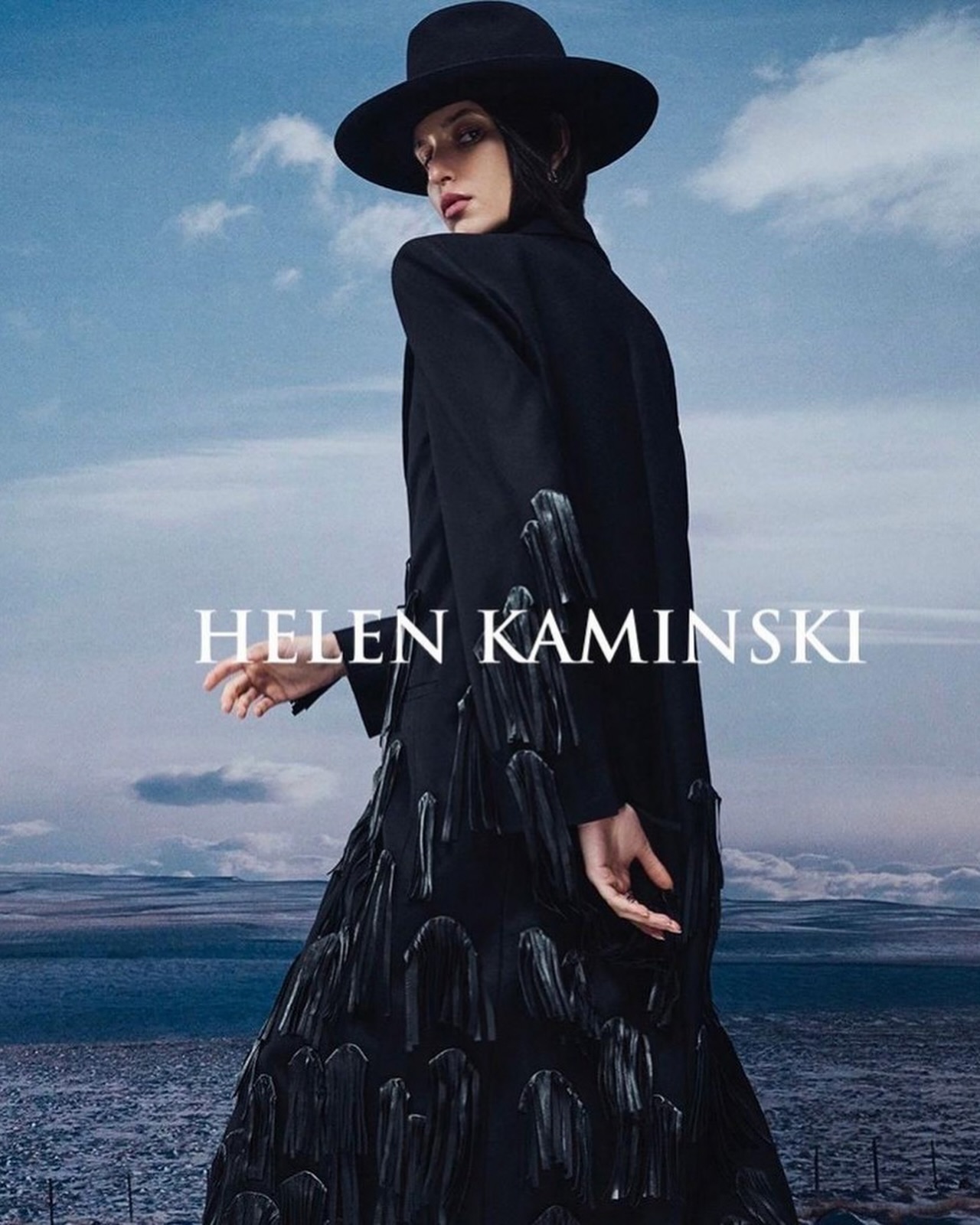 Model wearing a black hat and long black coat with fringe details against a landscape background with a cloudy sky.