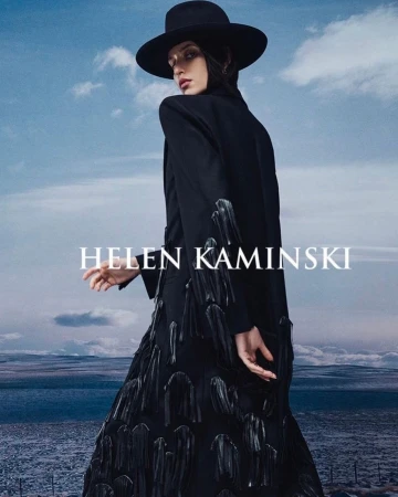 Model wearing a black hat and long black coat with fringe details against a landscape background with a cloudy sky.