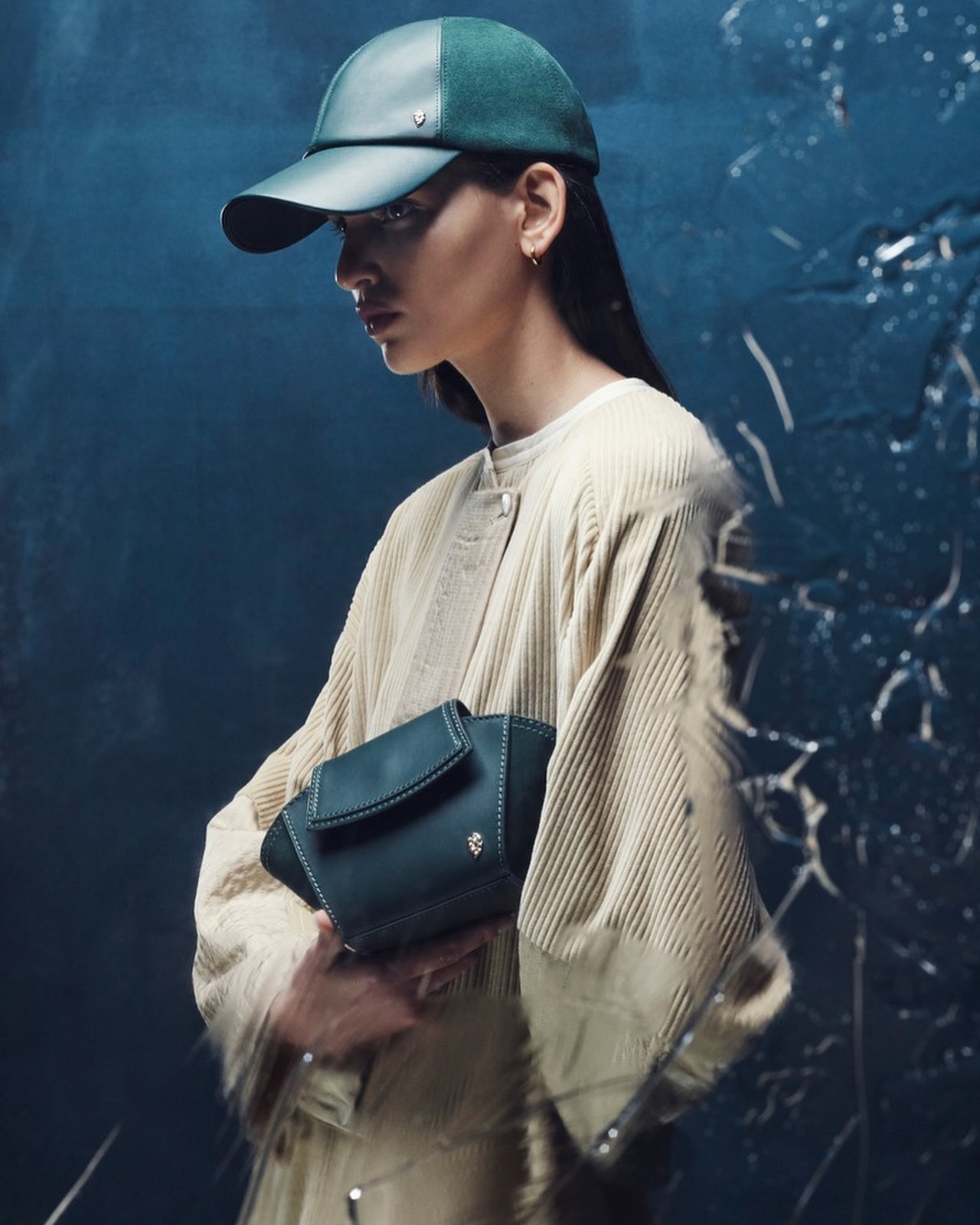 Woman wearing a green baseball cap and holding a dark green purse. She is wearing a corduroy coat in a studio with a textured backdrop.