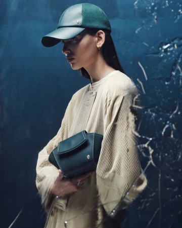 Woman wearing a green baseball cap and holding a dark green purse. She is wearing a corduroy coat in a studio with a textured backdrop.