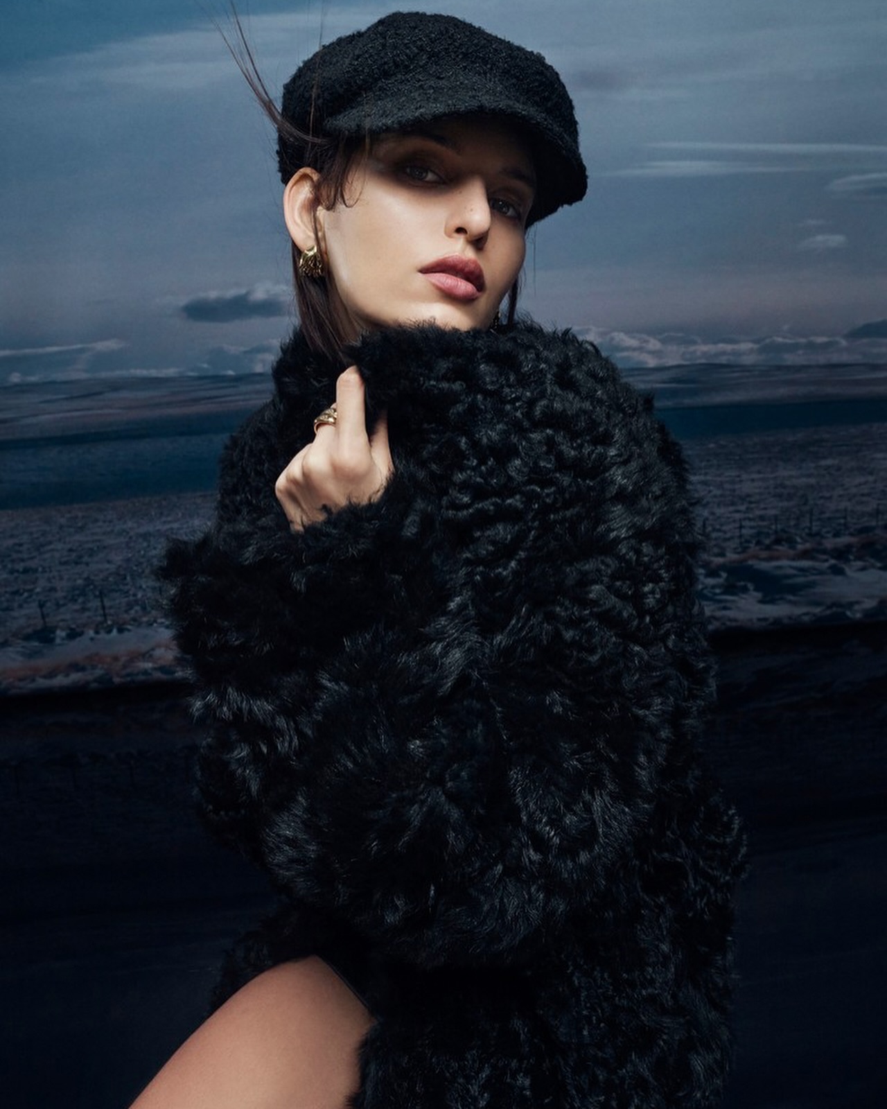 Fashion portrait of a woman with short hair wearing a black hat and fuzzy black coat posing against a background of a cloudy landscape.