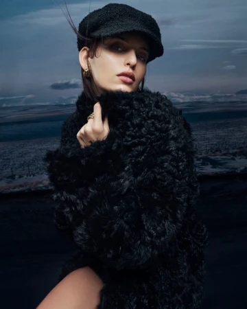 Fashion portrait of a woman with short hair wearing a black hat and fuzzy black coat posing against a background of a cloudy landscape.