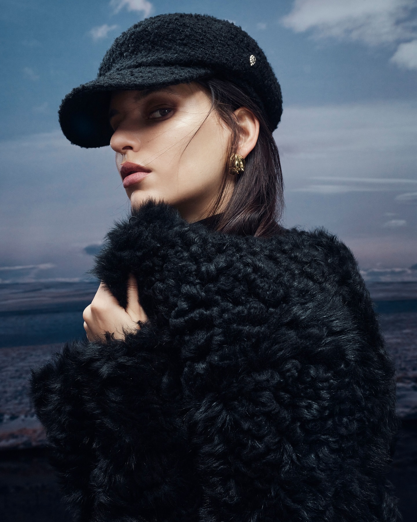 Model wearing a black textured cap and fluffy black coat against an overcast sky background for Helen Kaminski