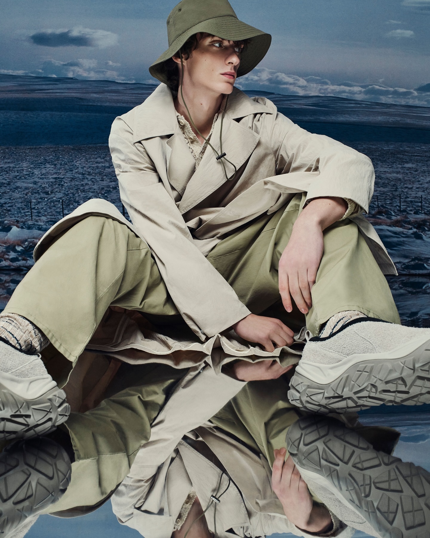 A young adult male sits outdoors wearing a bucket hat and a trench coat, with a mirror reflecting his image and a landscape in the background.