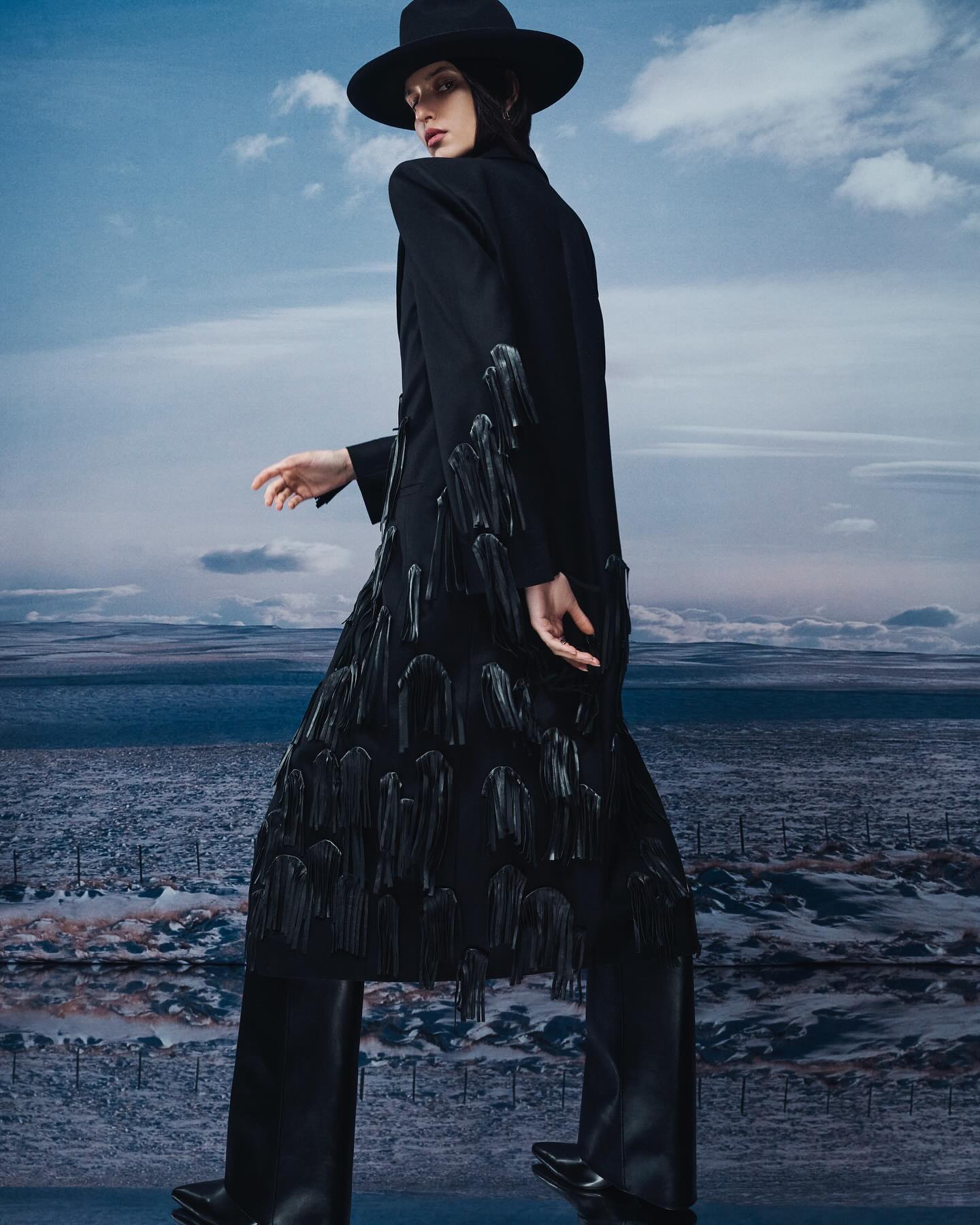 Model in a black hat and long black coat with fringe details standing against a dramatic cloudy sky and flat horizon.