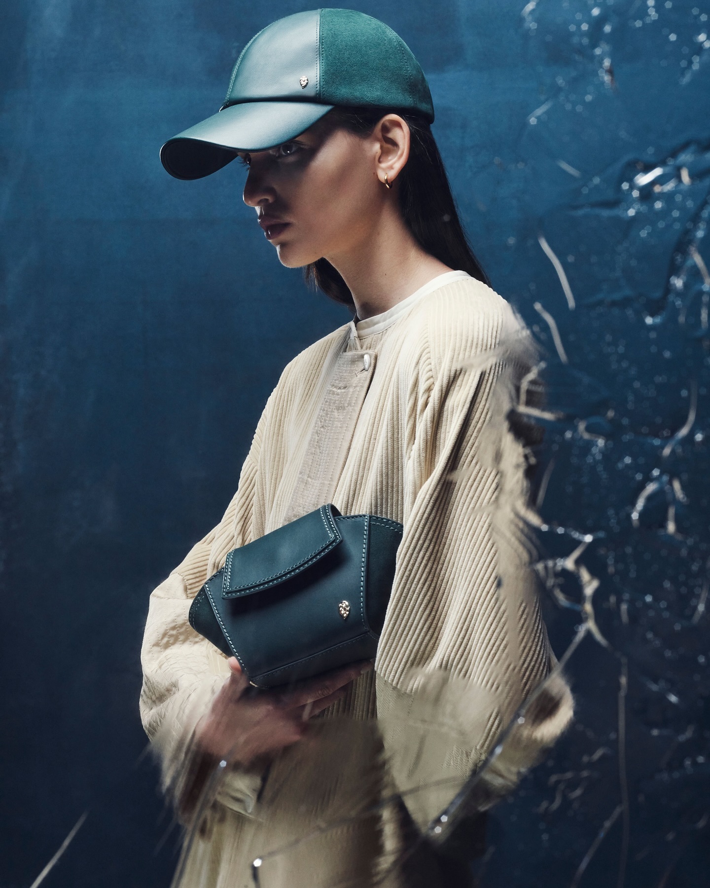 Woman in a green cap and beige corduroy jacket, holding a structured green bag. Soft studio lighting and a neutral-toned color palette.