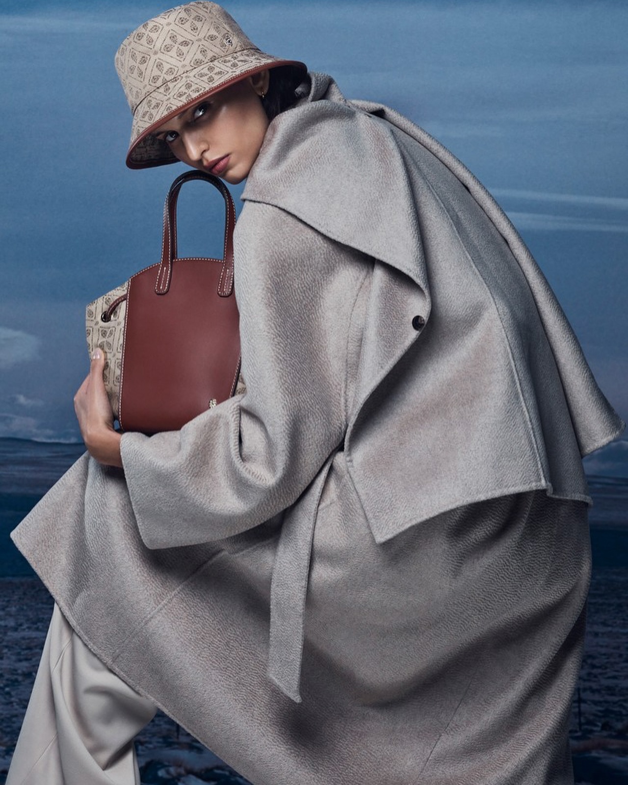 Model wearing a patterned bucket hat, grey coat and holding a structured brown handbag against a landscape sky background