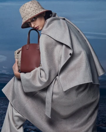 Model wearing a patterned bucket hat, grey coat and holding a structured brown handbag against a landscape sky background