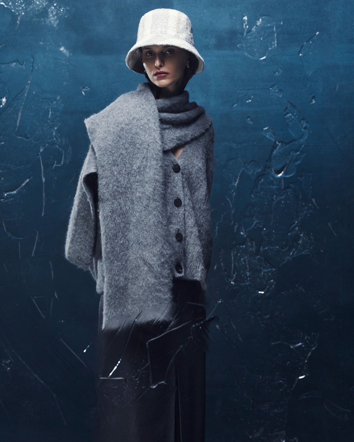 Woman in grey cardigan, scarf, and hat poses against a textured blue background. Fashion shot with natural light and soft focus.