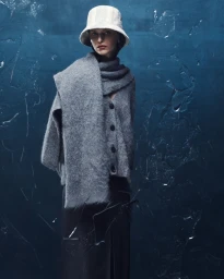 Model wearing a grey knit scarf, cardigan, and light bucket hat against a dark blue textured background.