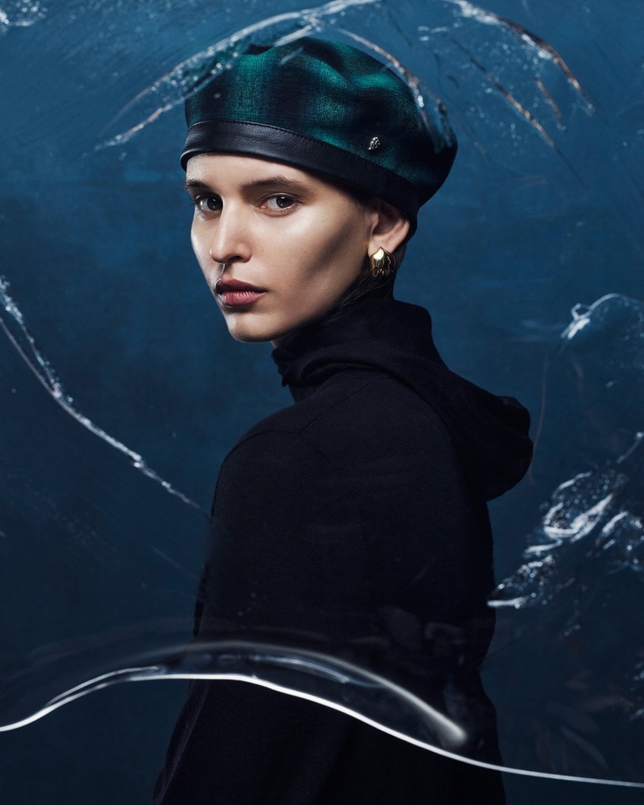 Portrait of a woman with fair skin wearing a black turtleneck and a green and blue plaid beret with a blue abstract background.