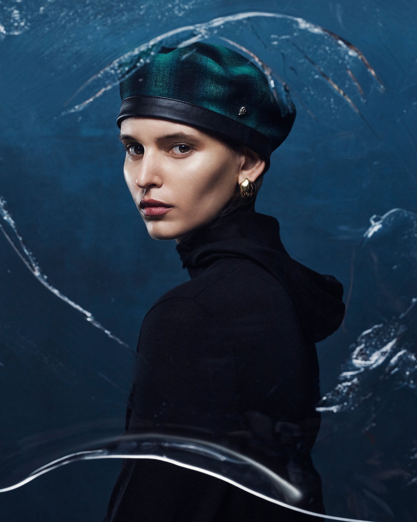 Woman in a green plaid beret and black turtleneck posing in a studio environment with a dark blue background and abstract white lines.
