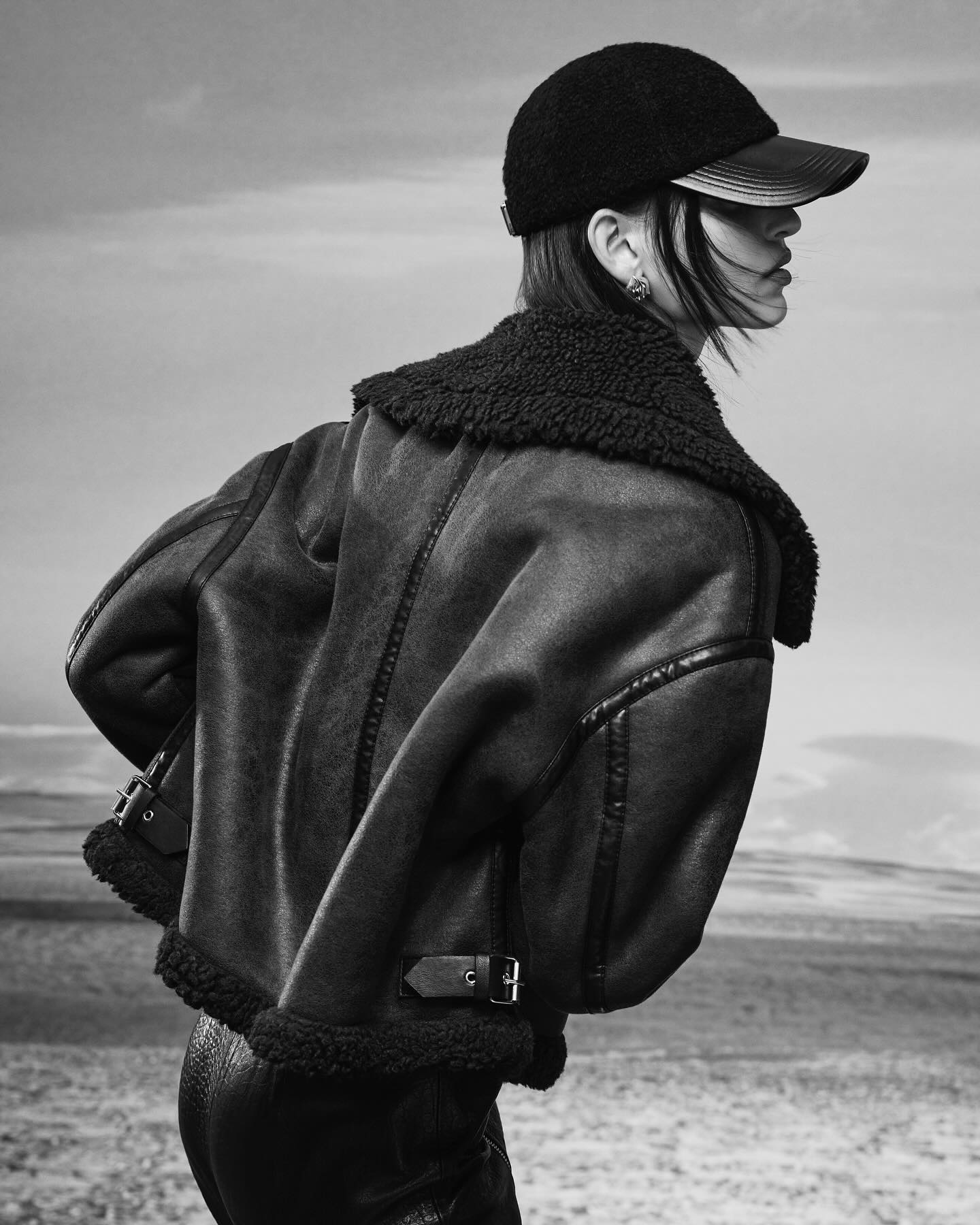 Black and white fashion shot of a woman in a shearling-lined aviator jacket and baseball cap standing in an open landscape.