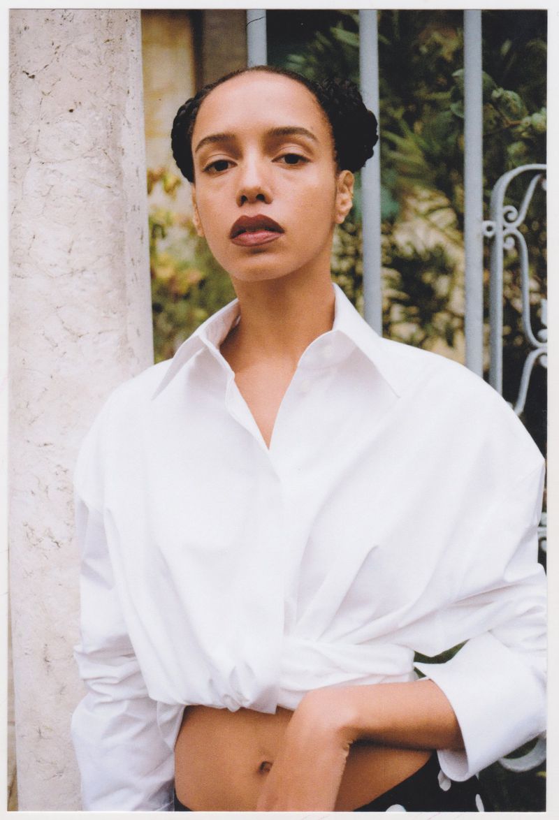 Portrait of a young adult woman with braided hair posing in a white shirt with a column and a metal gate in the background.