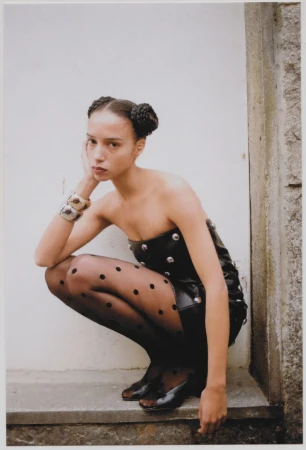 A young woman in a black strapless studded dress and polka dot tights crouches against a white wall, posing in a fashion shot.