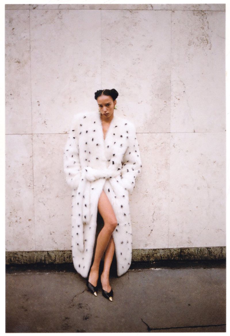 A woman in a long white fur coat with black polka dots poses against a textured wall in an outdoor setting with black shoes.