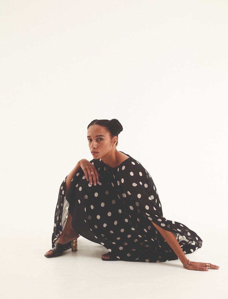 Woman in a polka dot dress poses while seated against a white background, looking directly at the camera in a studio shot.