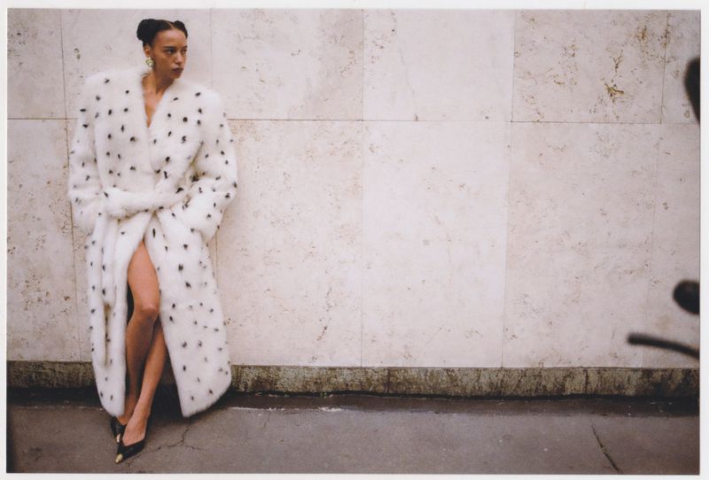 Elegant woman in a patterned white coat poses against a textured wall, showcasing fashion and style, highlighting her sophistication.