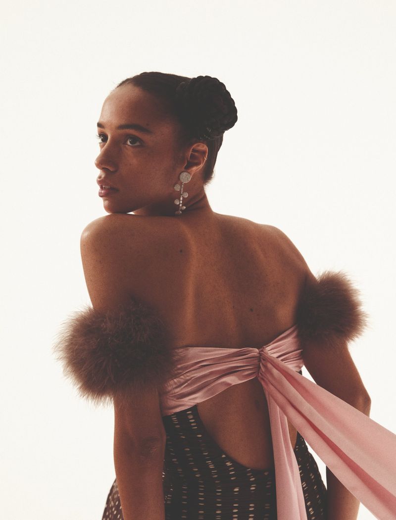 Elegant woman with dark hair in a high bun, wearing a dress with fur accents and statement earrings on a white background.