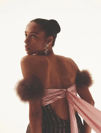Elegant woman with dark hair in a high bun, wearing a dress with fur accents and statement earrings on a white background.