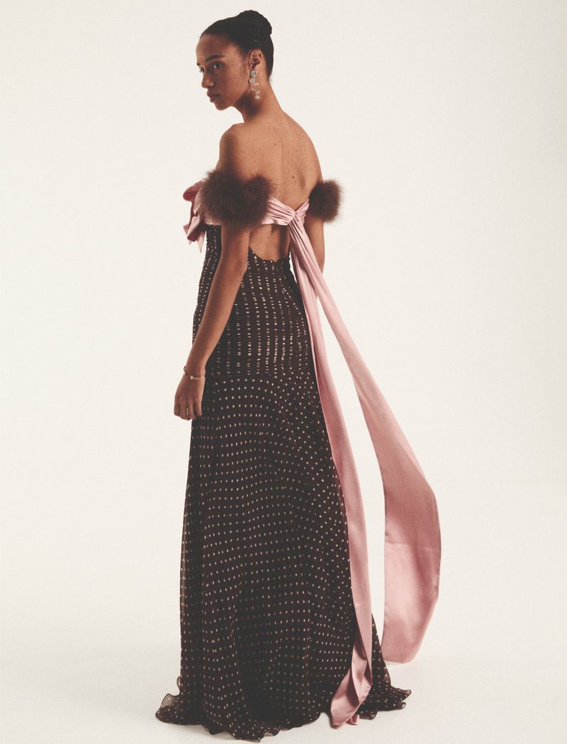 Back view of an adult female fashion model with dark hair, wearing a long brown and white dress with pink bow, looking over her shoulder.