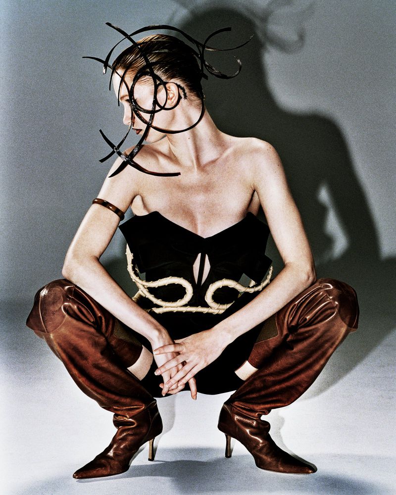 High-fashion portrait of a woman in a black strapless dress and thigh-high boots, kneeling with an artistic headpiece, in a studio.