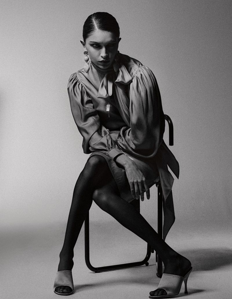 A woman sits on a black chair wearing a short-sleeve dress, sheer black tights, and heeled shoes against a neutral background.