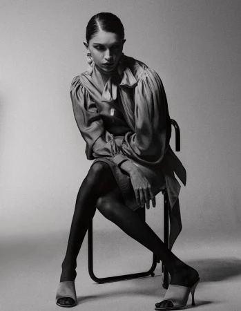 Monochrome portrait of a woman seated on a chair in a silk blouse, tights, and mule heels against a studio background.