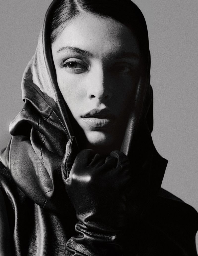 Monochrome portrait of a woman wearing a sculpted black leather hood and leather glove against a studio background.