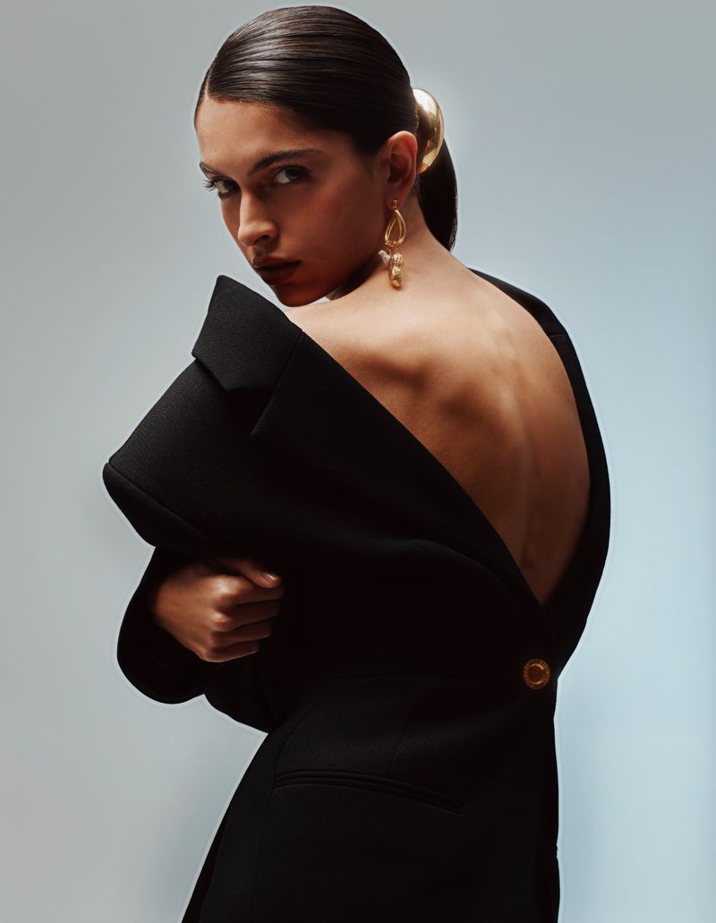 A woman in a black off-the-shoulder blazer poses in a studio, looking over her shoulder with gold drop earrings and hair accessory.