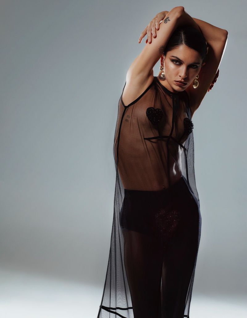 Brunette adult female in a sheer black dress strikes a fashion pose in a studio, showcasing contemporary style, earrings, and tattoo art.