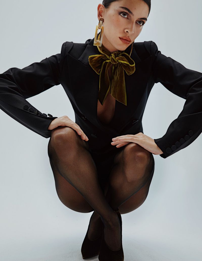 Elegant woman in a black suit and sheer tights posing in a photo studio with minimalist styling and soft lighting.
