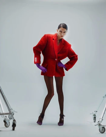 Model posing in an oversized red blazer, purple gloves, and sheer tights on a light grey studio sweep with equipment wheels visible.