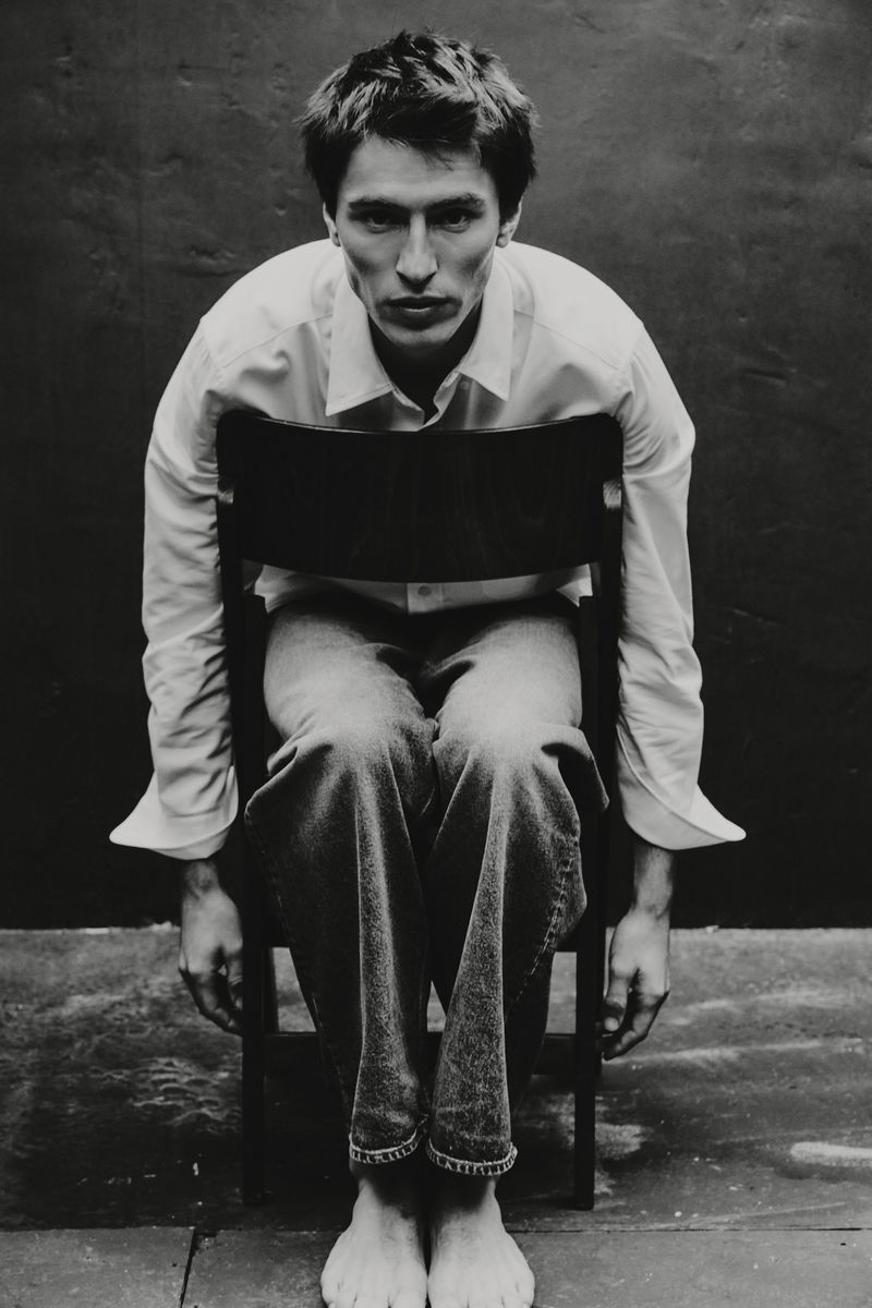 Model Clement Laguardia sits backwards on a wooden chair wearing a white shirt and cuffed jeans in front of a dark backdrop.