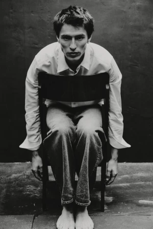 Model Clement Laguardia sits backwards on a wooden chair wearing a white shirt and cuffed jeans in front of a dark backdrop.