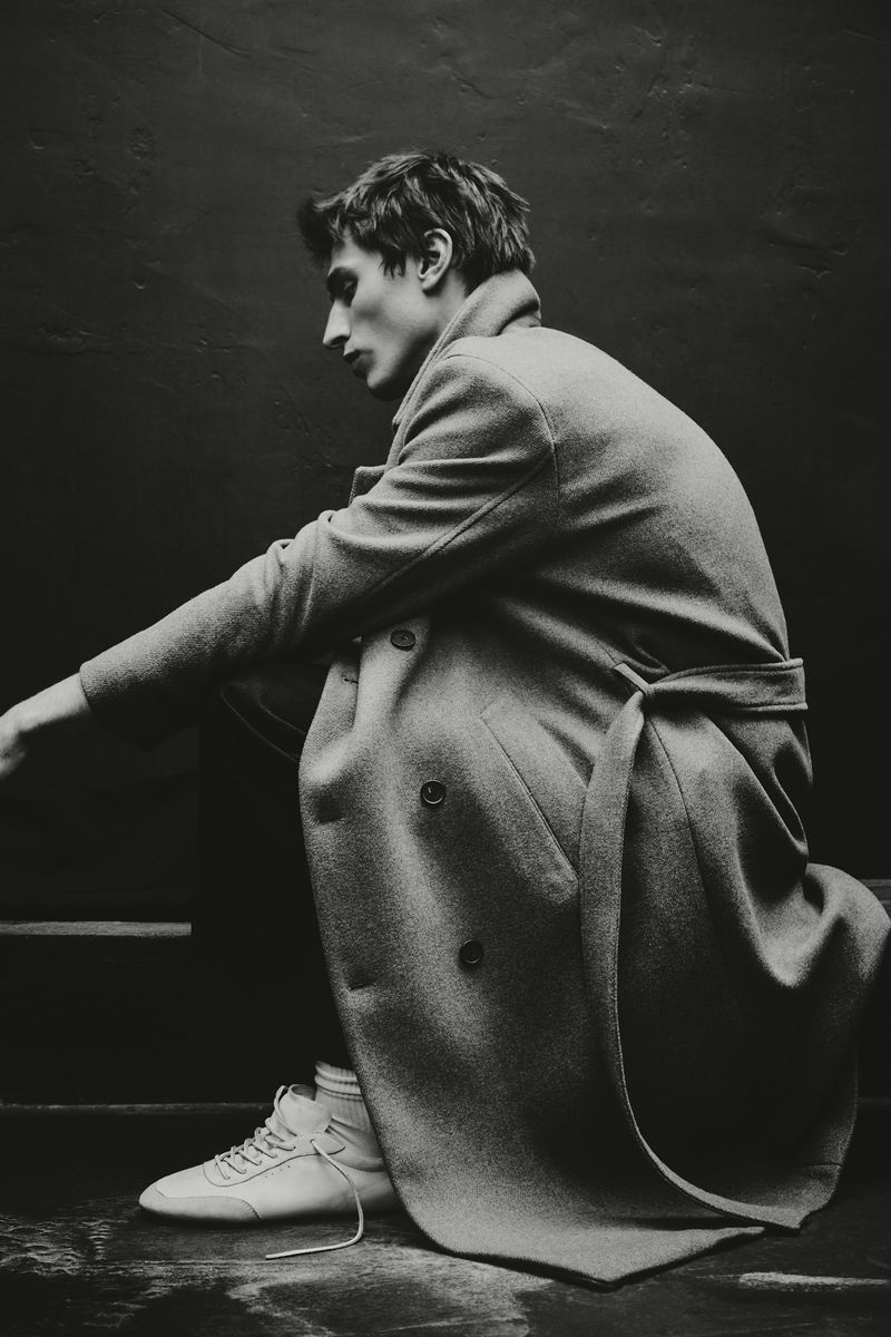 Monochrome fashion portrait of a man in a trench coat and sneakers, captured in a squatting pose against a dark wall.